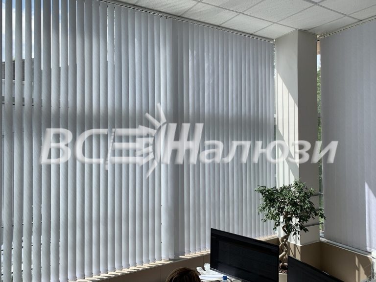 Белые вертикальные жалюзи вдоль угловых окон офиса