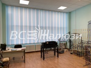 Голубые полосатые вертикальные жалюзи вдоль широкого окна светлого кабинета