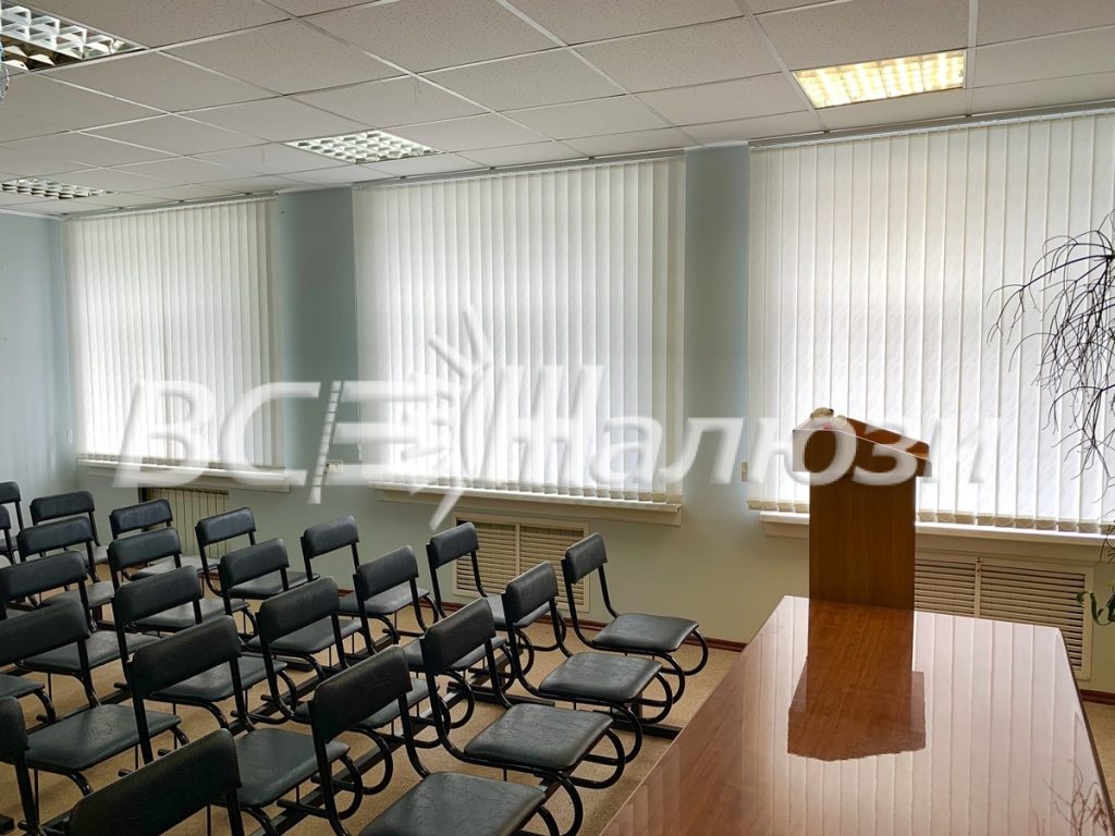 Светлые вертикальные жалюзи в зале с рядами стульев и трибуной