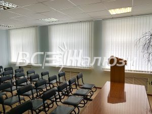 Светлые вертикальные жалюзи в зале с рядами стульев и трибуной