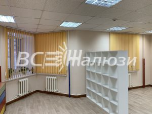 Жёлтые вертикальные жалюзи на окнах комнаты с белыми стеллажами