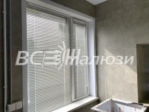 Белые горизонтальные жалюзи на окне комнаты с серыми стенами