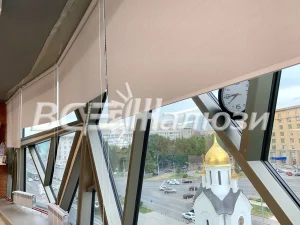 Бежевые рулонные шторы приспущены на панорамном остеклении с видом на город