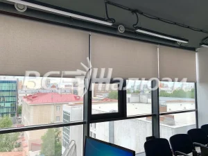 Серые рулонные шторы наполовину опущены на окнах офиса