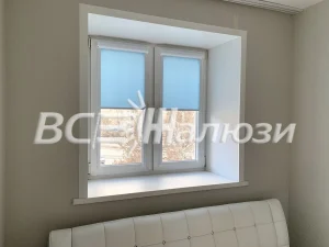 Голубые рулонные шторы подняты на небольшом окне светлой комнаты