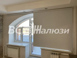 Бежевая рулонная штора закрывает балконный блок рядом с арочным окном