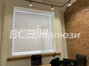 Белая рулонная штора на широком окне офиса с кирпичной стеной