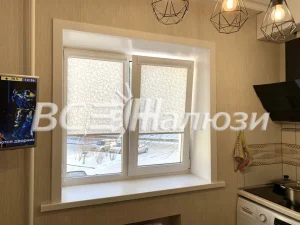 Светлые рулонные шторы подняты на кухонном окне, за стеклом двор
