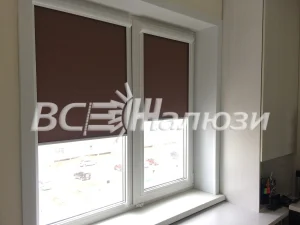 Шоколадные рулонные шторы наполовину опущены на белом пластиковом окне