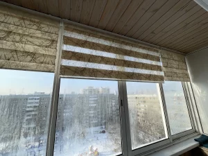 Бежевая штора зебра приспущена на окне балкона с деревянным потолком