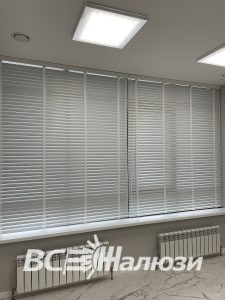 Белые жалюзи полностью закрыты на широком окне светлого кабинета
